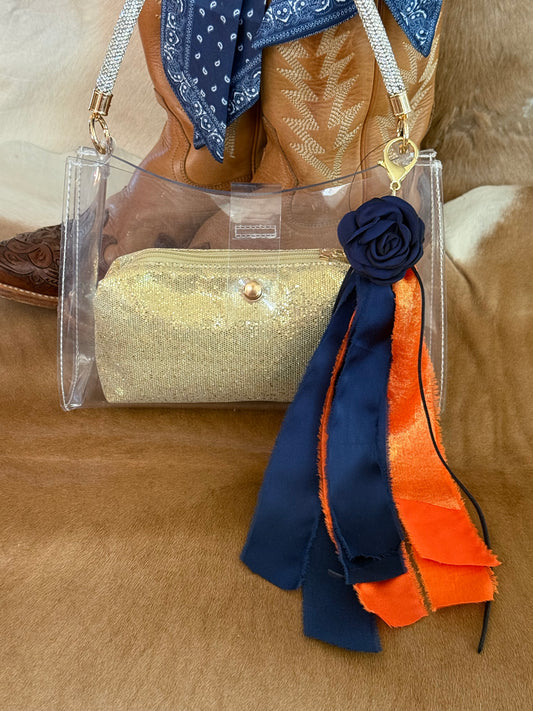 A clear acrylic shoulder bag with a gold glitter pouch inside, styled with a navy blue fabric rose and navy/orange ribbon charm on the front. The bag is shown with western boots and a blue bandana in the background on a brown hide surface.